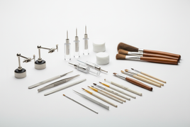 Precision modeling tools including glue applicators, magnetic holders, cleaning discs and brush dusters arranged on white workbench for scale model assembly