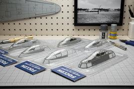 Model airplane canopies in plastic packaging on a hobby workbench, tools and paint nearby.