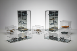 Assorted clear acrylic and glass display cases of various sizes arranged on a light gray surface, some containing scale model cars and aircraft, for protecting and showcasing hobby collectibles.