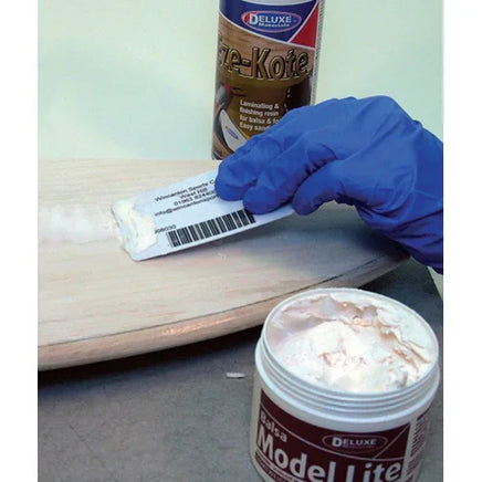 Person applying a white substance from a tube labeled 'Model Lite' to a wooden surface, with a can of 'Deluxe Fire-Kote' in the background.