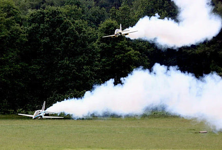 Two airplanes performing aerial stunts with smoke trails over a grassy field with trees in the background.