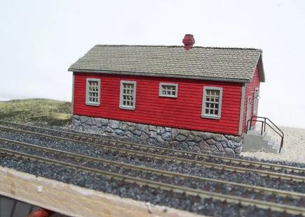 Model of a red building with gray roof and stone foundation on a model railway track
