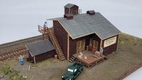 Model of a rustic building with a green truck in front on a plain background