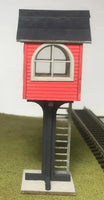 Model of a small red building with a black roof on a stand, near a train track.