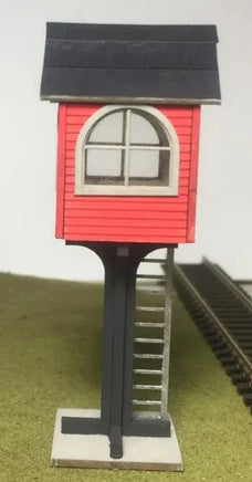 Model of a small red building with a black roof on a stand, near a train track.