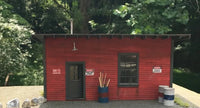 Small red building with a door and window, surrounded by trees and barrels.