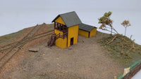 Model of a yellow building with a gray roof on a miniature landscape with tracks and trees.