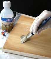 Person applying a wood finish to a wooden surface with a brush, next to a bottle of Eze-Kote.