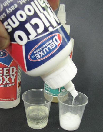 Two plastic cups with a bottle of Deluxe Speed Glue being poured into them on a dark background.