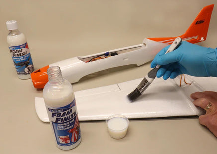Person applying foam finish to a model airplane on a table with Gloss Finish bottles and a brush.