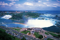 Aerial view of Niagara Falls with a rainbow, cars on the road below, and buildings in the distance.