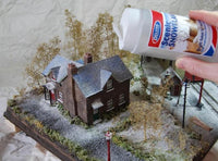 Model train layout with a house and trees, being dusted with scenic snow by a hand holding a can of snow.