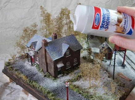 Model train layout with a house and trees, being dusted with scenic snow by a hand holding a can of snow.