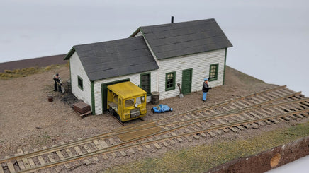Model train setup with a small building, yellow truck, and figures on a track.