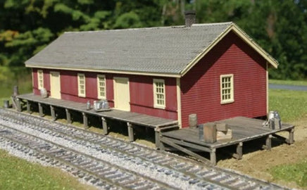 Model train station building with red exterior and wooden platform on a grassy area with tracks.