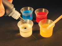 Person using a dropper to add liquid from a bottle into four small cups with different colored liquids on a dark surface.