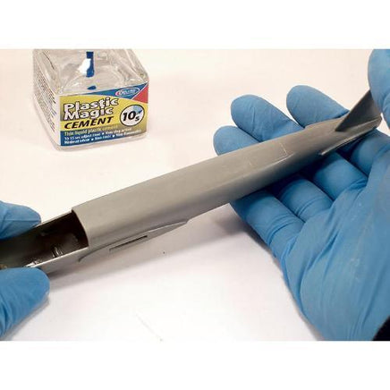 Person wearing blue gloves holding a metal object with Plastic Magic Cement box in the background.