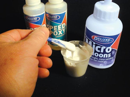 Hand holding a small container of white material with glue bottles labeled 'Deluxe' in the background.