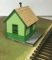 Model train station building with green siding and brown roof next to train tracks.