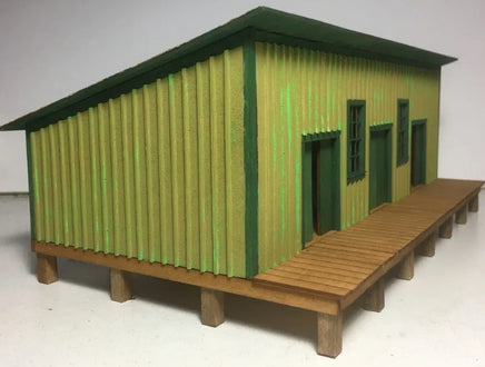 Model of a small building with green corrugated metal siding and a wooden deck on a white background