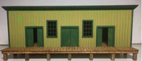 Model of a small building with green doors and windows on a wooden platform.
