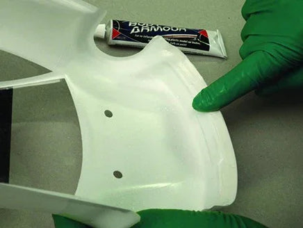 White plastic part with holes, green gloved hand pointing, and a tube of Bostik Damaud on a gray background.