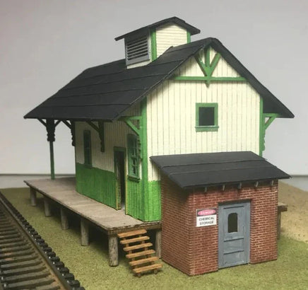 Model train station with green and white building and brick storage shed on a plain background