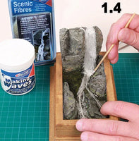 Person working on a miniature landscape with rocks and fibers, using a container labeled 'Waking Waves' and a box of 'Scenic Fibres'.
