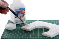 Person applying foam putty to a model on a cutting mat with a bottle of foam putty in the background.